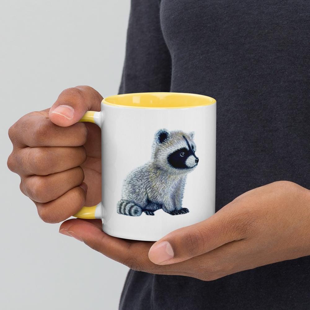 Yellow ceramic mug with Be You in bold text above a detailed baby raccoon illustration, featuring fine textures and lifelike details. Glossy finish with a standard-sized handle.