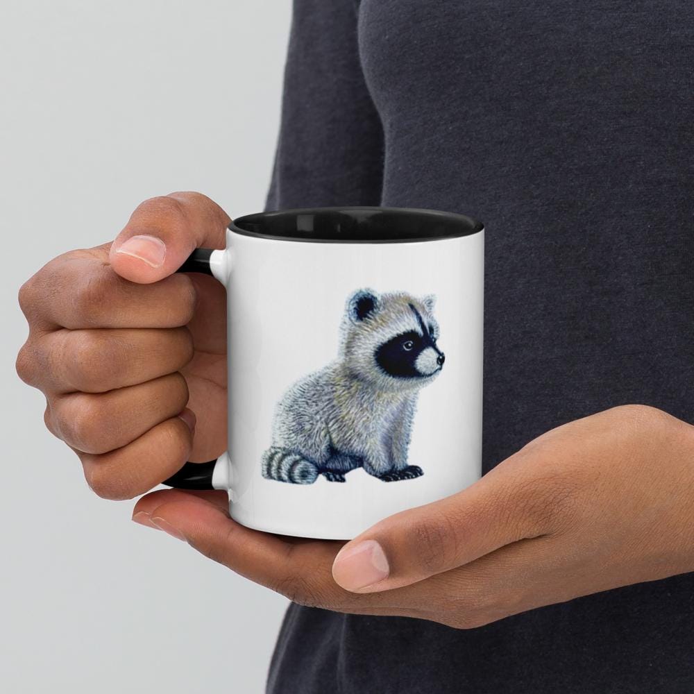 Black ceramic mug with Be You in bold text above a detailed baby raccoon illustration, featuring fine textures and lifelike details. Glossy finish with a standard-sized handle.