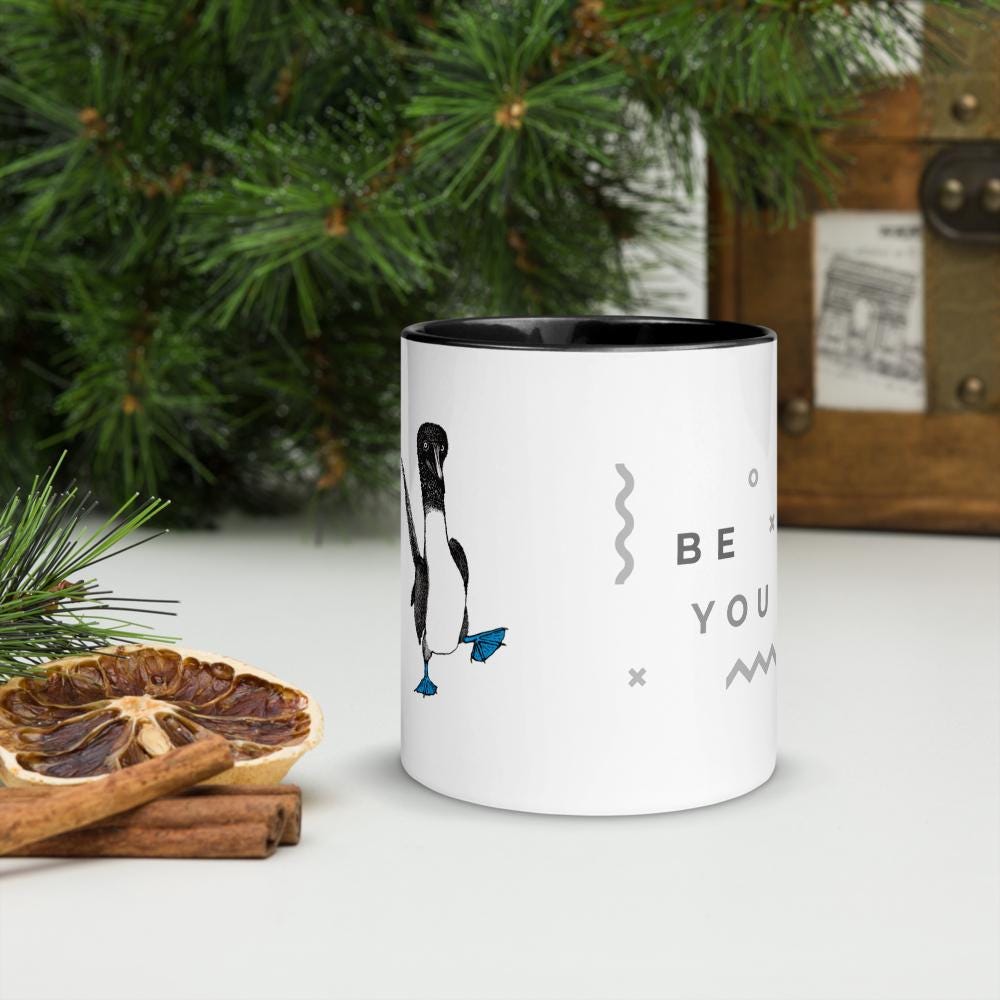 Black ceramic mug with Be You in bold text above a detailed illustration of a blue-footed booby. Features fine textures and lifelike details. Glossy finish with a standard-sized handle.