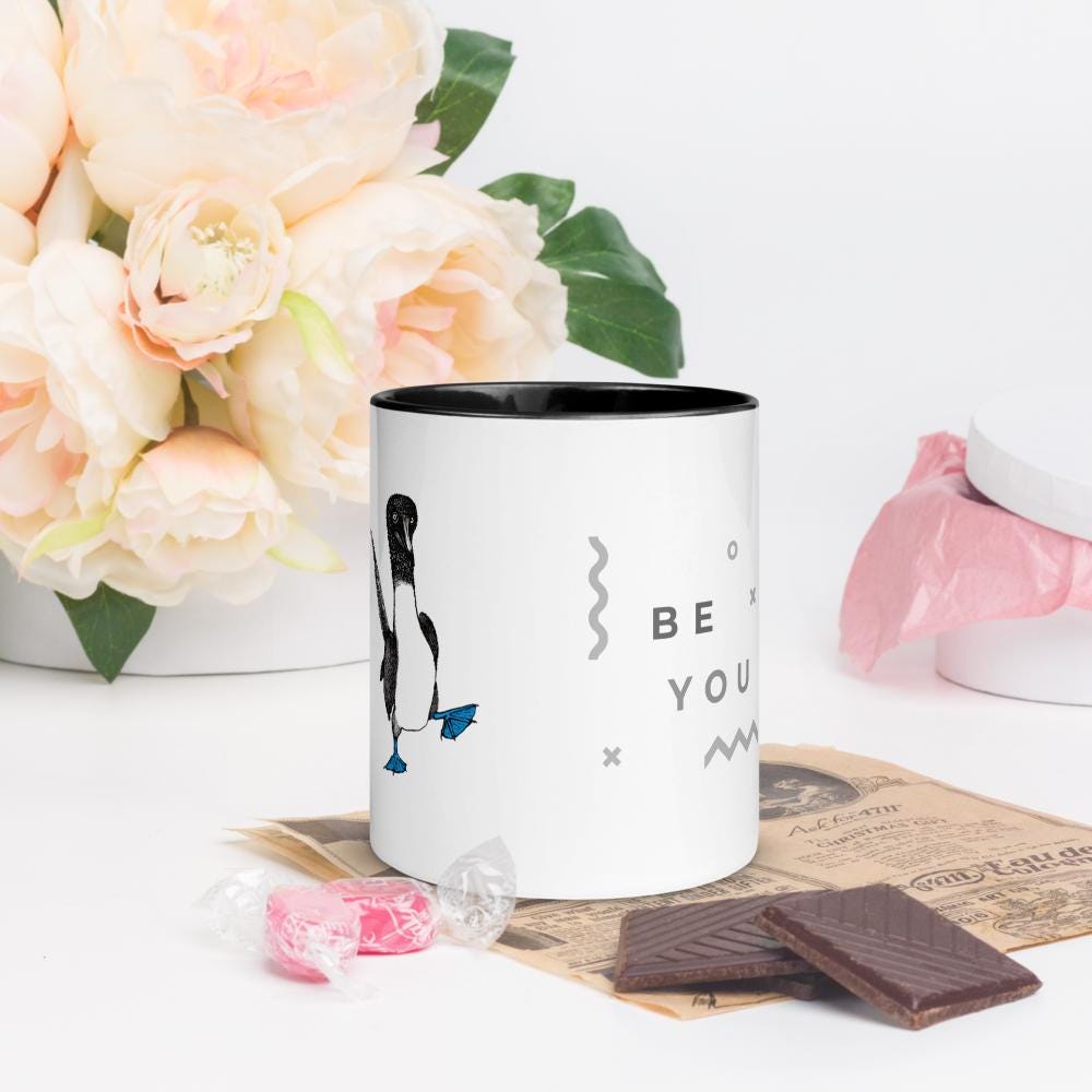 Black ceramic mug with Be You in bold text above a detailed illustration of a blue-footed booby. Features fine textures and lifelike details. Glossy finish with a standard-sized handle.