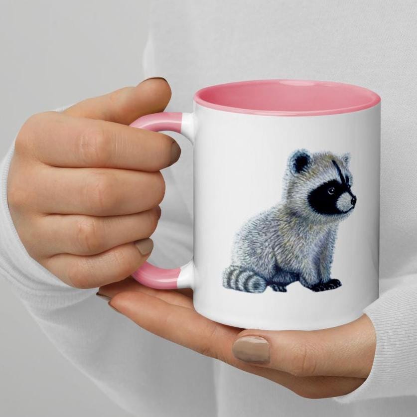 Pink ceramic mug featuring a detailed illustration of a baby raccoon, drawn with fine, realistic textures. The raccoon is positioned on the front of the mug. Smooth glossy finish with a standard-sized handle for comfortable grip.